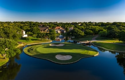 A golf green completely surrounded by water with homes in the background.