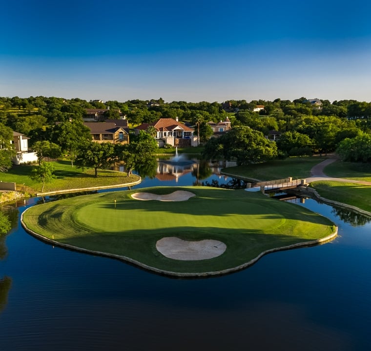 A golf green completely surrounded by water with homes in the background.