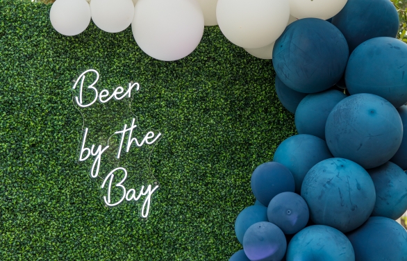 white and blue balloons on a large hedge next to light up letters saying beer by the bay