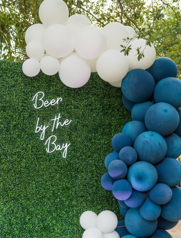 white and blue balloons on a large hedge next to light up letters saying beer by the bay