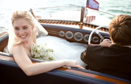 Newlyweds leaving ceremony on wooden boat, bride is smiling and looking back at her family.
