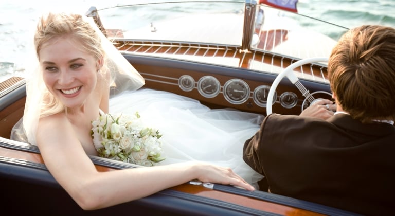 Newlyweds leaving ceremony on wooden boat, bride is smiling and looking back at her family.