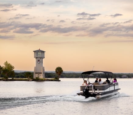 Pontoon boat driving away from the camera into the sunset next to the lighthouse on the lake.