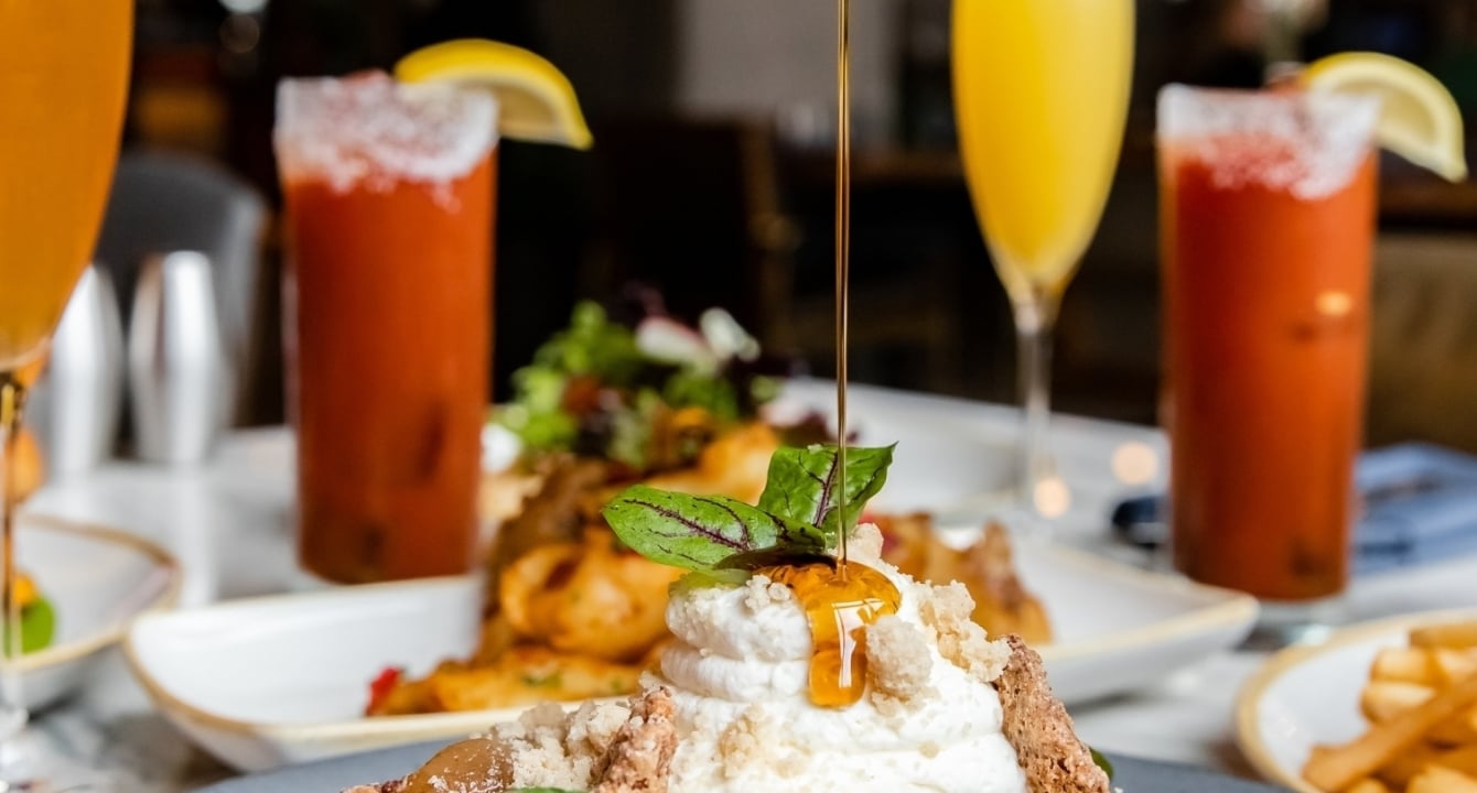 Maple syurp being poured by a server- just the hand is in the shot- on a chicken and waffles dish. With bloody marys and mimosas in the background